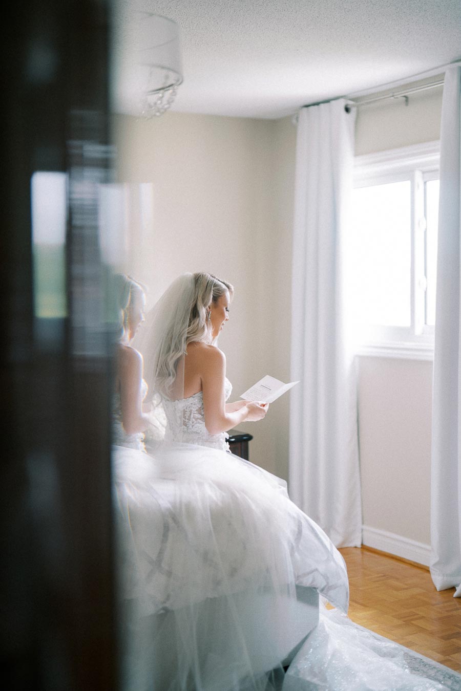 A bride in a white wedding dress and veil sitting by a window, reading a letter with a soft smile on her face, surrounded by natural light and delicate curtains, creating a serene and intimate atmosphere.