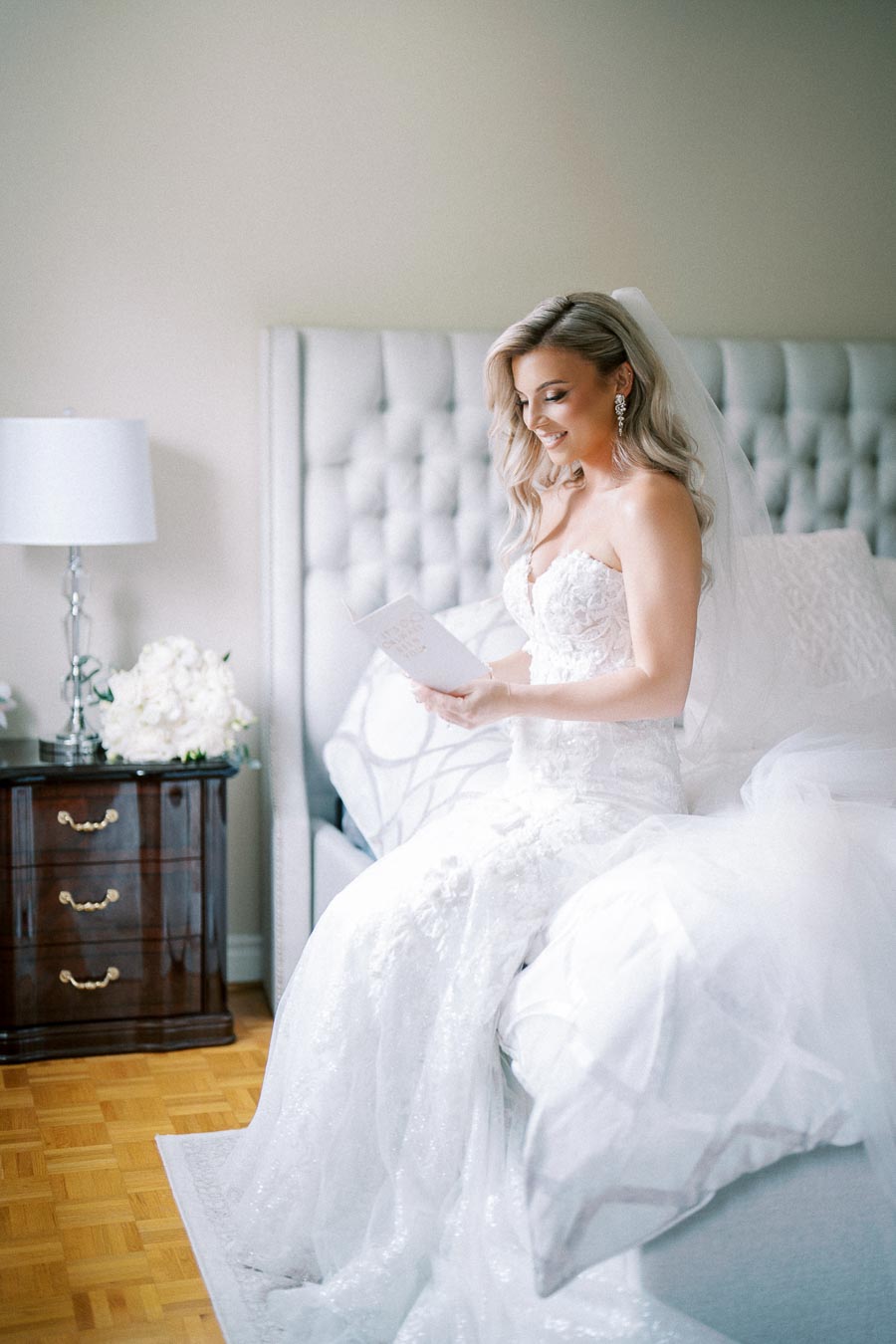 Bride in a white wedding dress sitting on a bed, reading a letter with a joyful expression, in a softly lit bedroom setting.