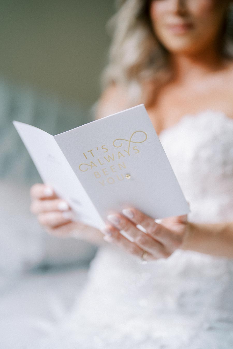 A bride in a white wedding dress holding a card with the words It's Always Been You in gold lettering, conveying a romantic and heartfelt wedding moment.