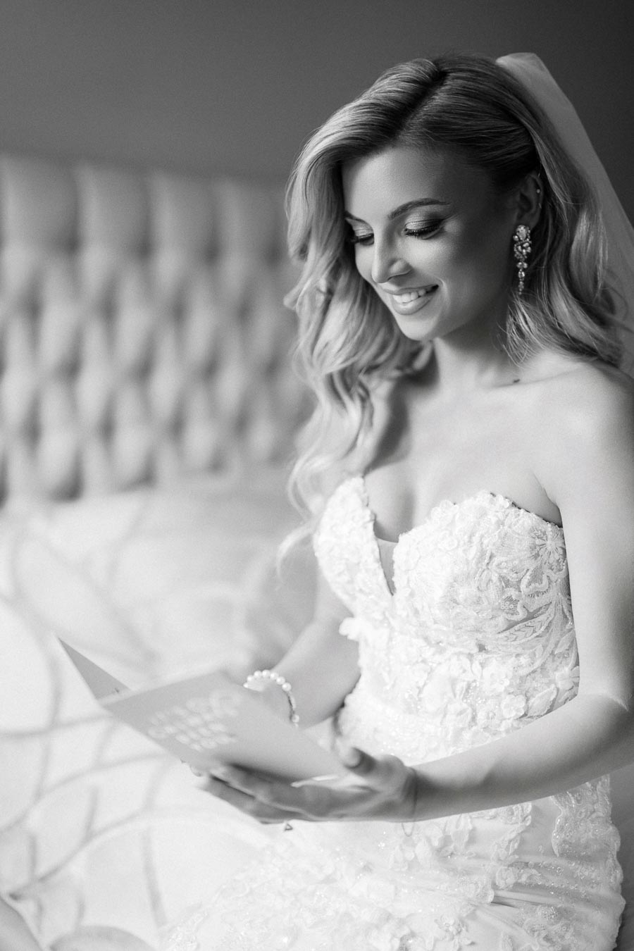 Black and white photo of a smiling bride in an elegant lace wedding gown holding a card, sitting on a bed with tufted headboard.