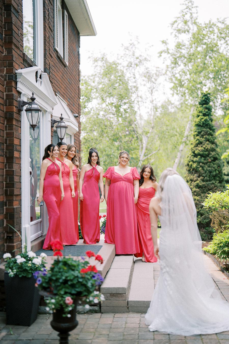Bridesmaids in vibrant pink dresses stand on a brick house porch, eagerly watching the bride in a white gown approach on her wedding day, surrounded by lush greenery and flowers.