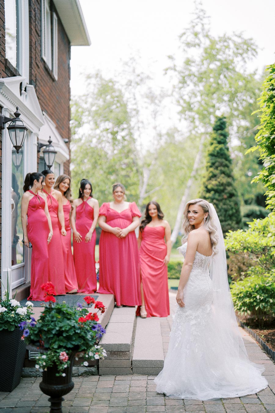 Bride in elegant white gown smiling at six bridesmaids in pink dresses, standing by a brick house surrounded by greenery and flowers.