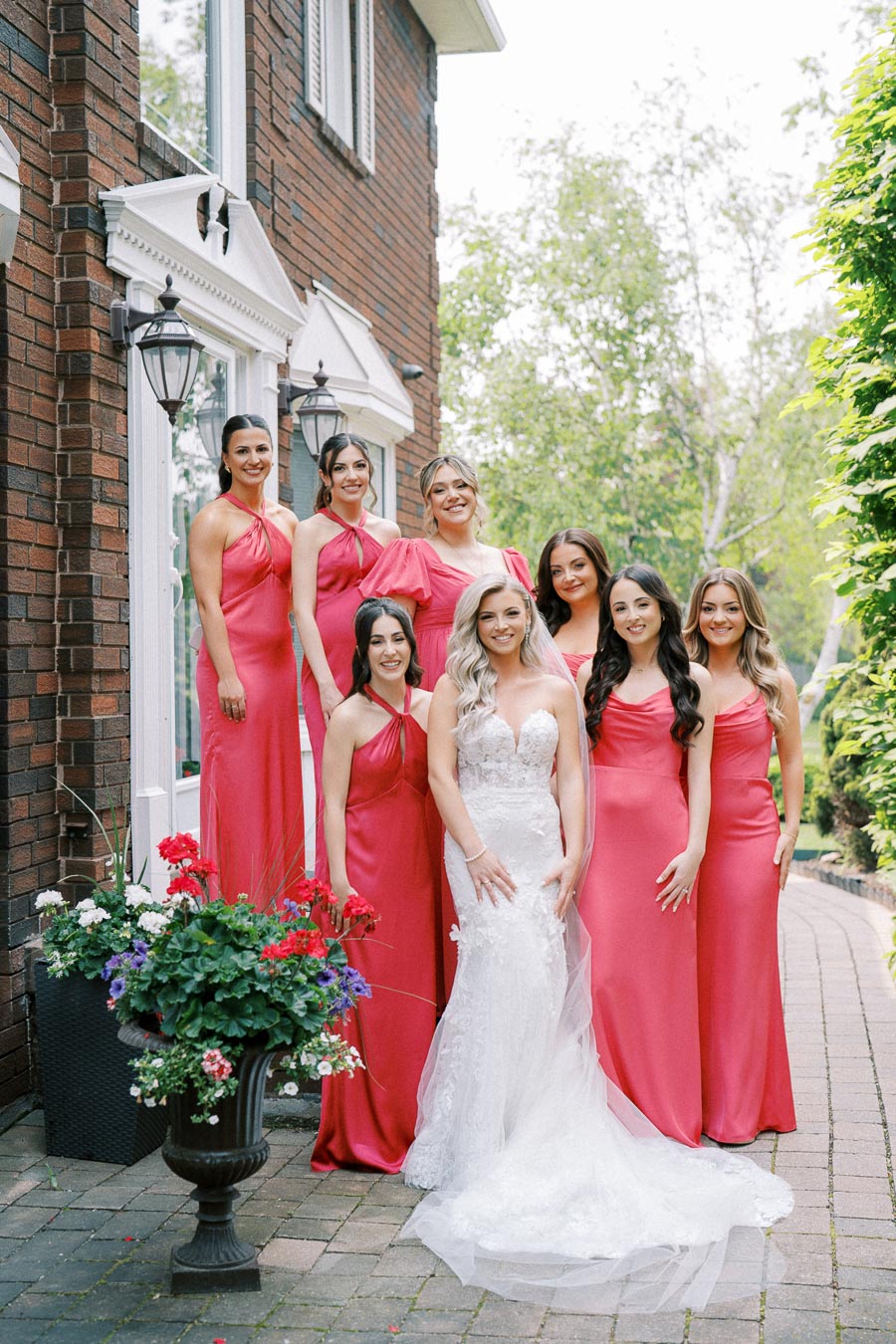 Bridal party photo featuring a bride in a white lace wedding dress surrounded by bridesmaids in vibrant pink dresses standing on a brick pathway outside a house with flowering plants nearby.