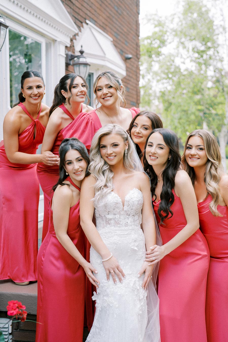 A joyful bride in a lace wedding gown surrounded by her bridesmaids wearing vibrant pink dresses, posing for a group photo outdoors in front of a brick building.