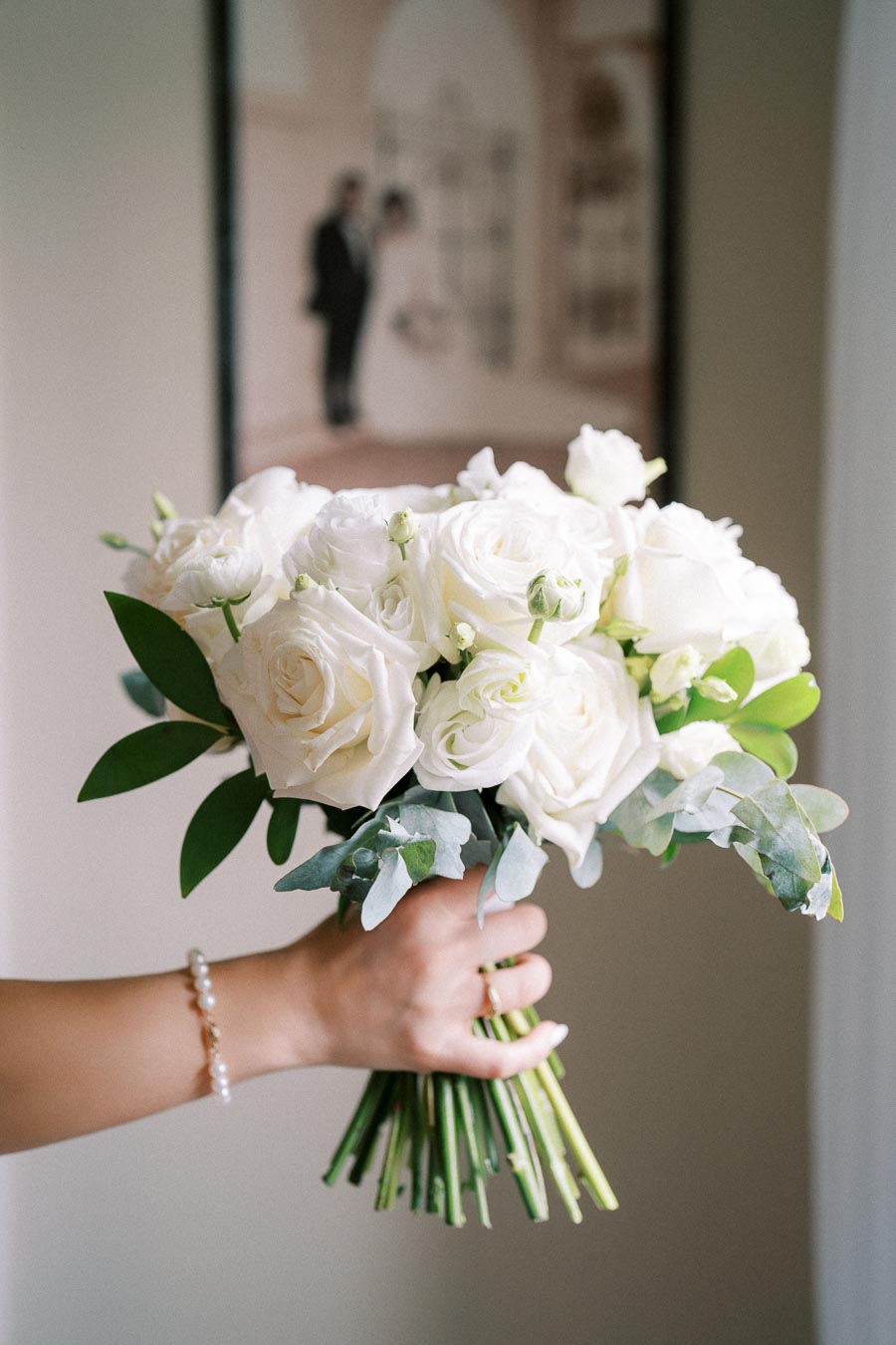 A hand holding a bouquet of white roses and greenery, with a blurred background of a romantic framed photo.