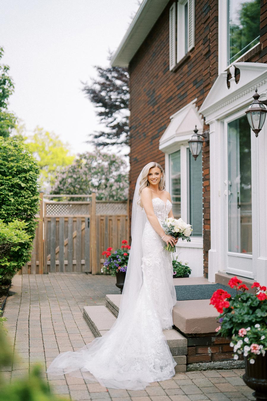 A bride in a white lace wedding gown stands outside a brick house, holding a bouquet of white flowers. She smiles with long blonde hair under a veil. Colorful flowers decorate the pathway, creating a vibrant garden setting.