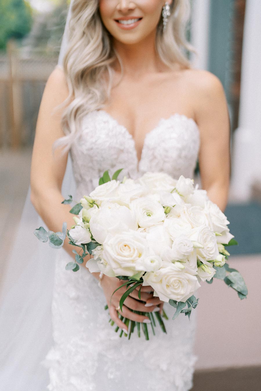 Bride in lace wedding dress holding a bouquet of white roses and greenery, smiling softly in an outdoor setting.
