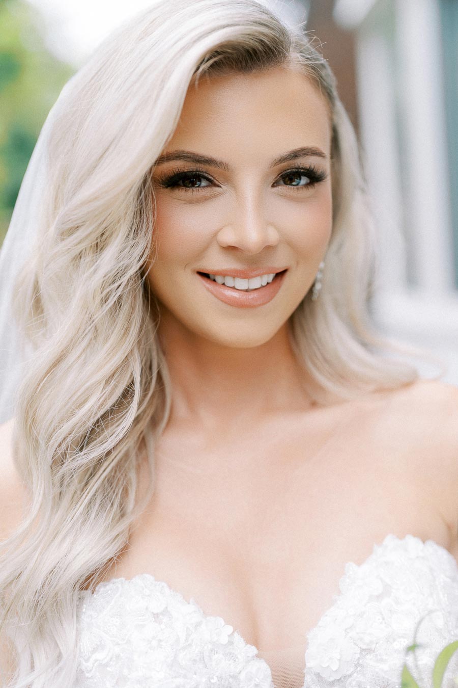 A bride with long, wavy blonde hair smiles on her wedding day, wearing a strapless white dress.