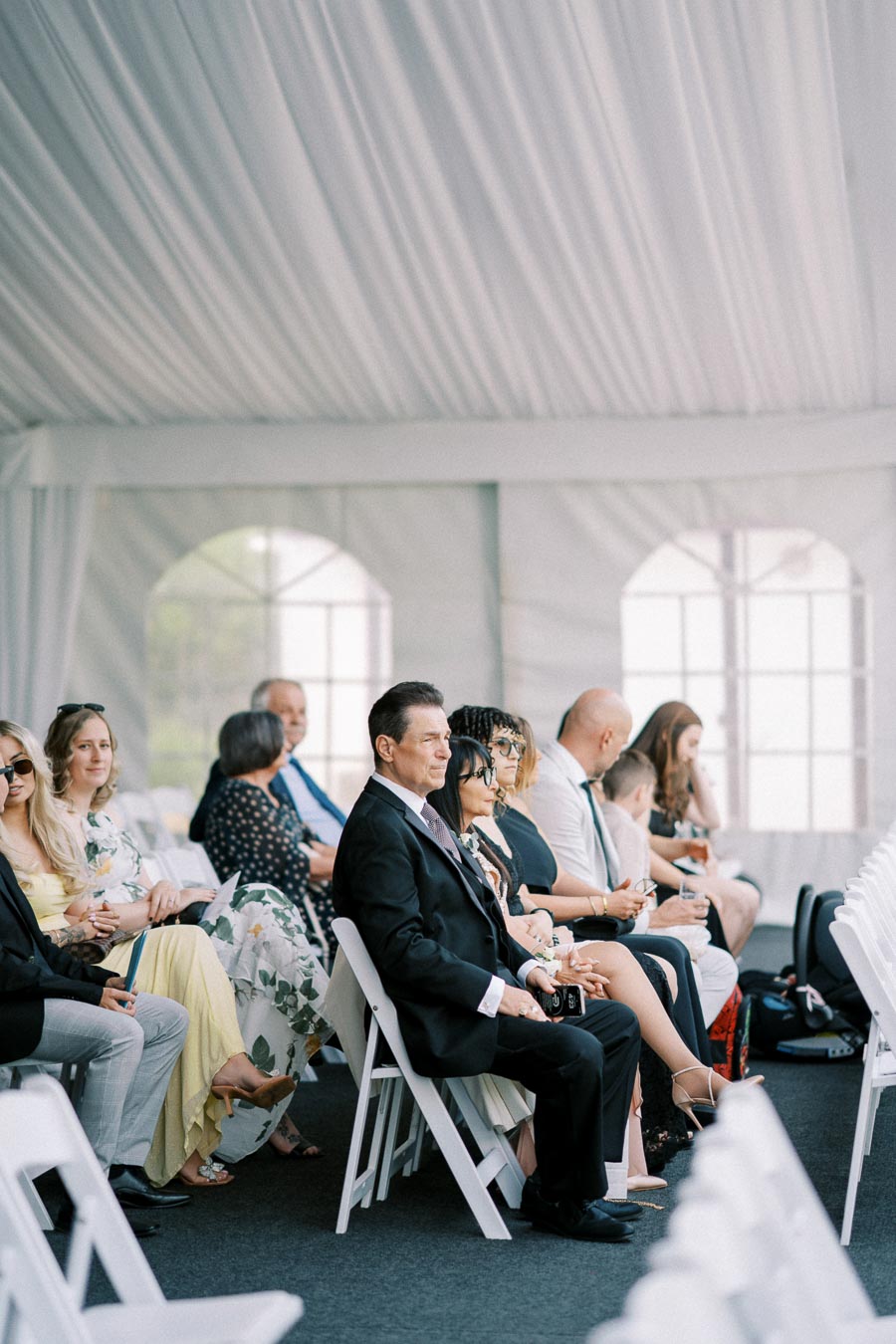 Guests seated in a white-tented venue for a formal event, wearing elegant attire, with attention focused forward.