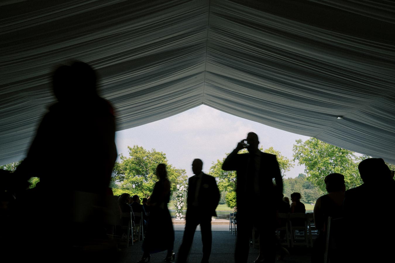 Silhouetted people walking under a large, elegant tent with natural light filtering in, greenery visible outside, creating a serene outdoor event setting.