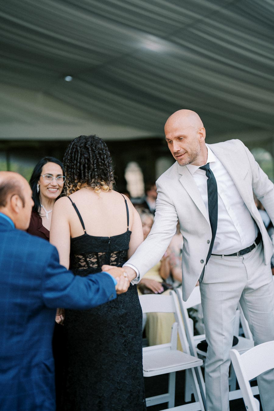 A man in a light gray suit shakes hands with a woman in a black lace dress during a formal event under a tent. Other guests are seated in the background.