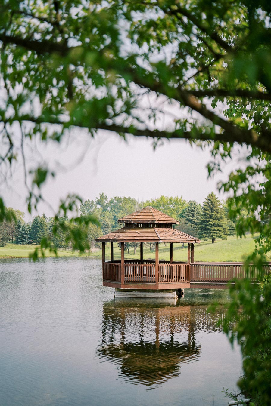 Scenic view of a wooden gazebo over a tranquil lake surrounded by lush green trees, with clear reflections in the water and a peaceful, natural setting.