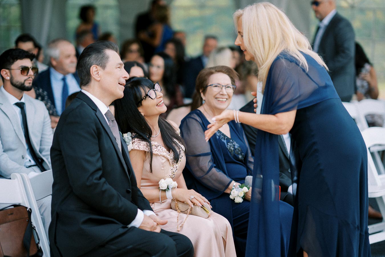 A group of elegant guests engages in conversation at a formal event, dressed in suits and evening gowns, with white chairs and a crowd in the background.