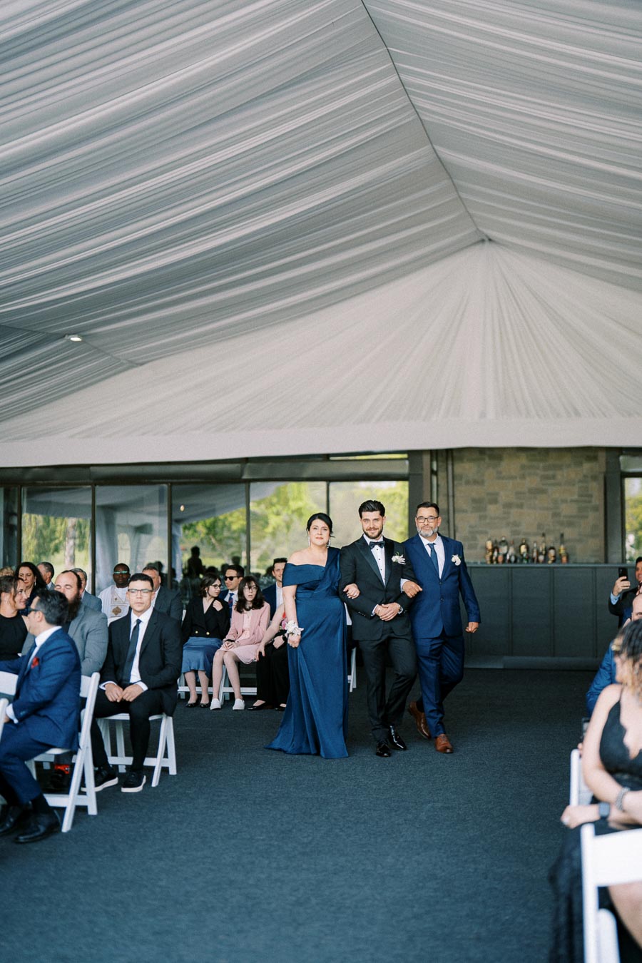 Elegant wedding procession under a white draped canopy, with a couple in formal attire leading guests through an aisle.