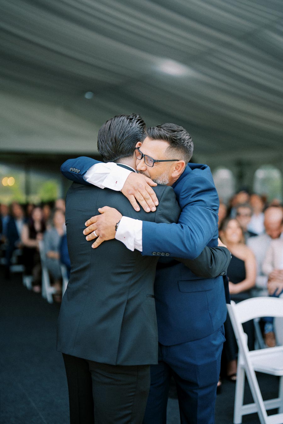 Two men in suits hugging at a formal event, surrounded by seated guests in an elegant venue.