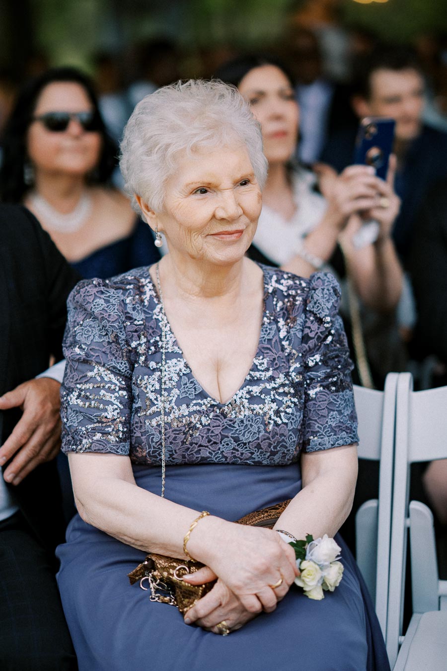 Elderly woman in a sequined lace dress seated outdoors at a formal event, holding a small clutch and a corsage, with attendees in the background.