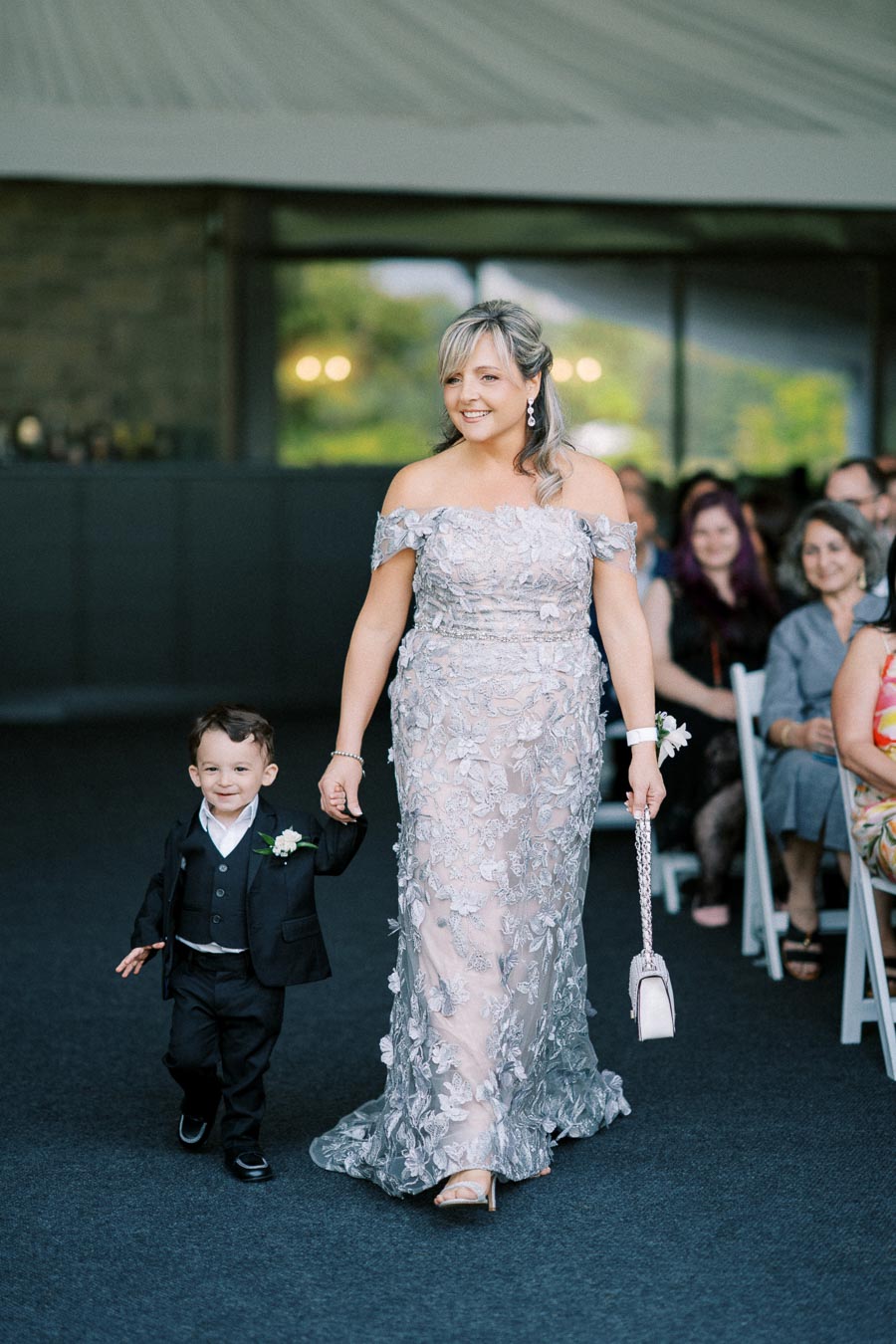 A woman in an elegant, off-the-shoulder floral gown walks down an aisle holding hands with a young boy in a black suit, surrounded by seated guests at a formal event.