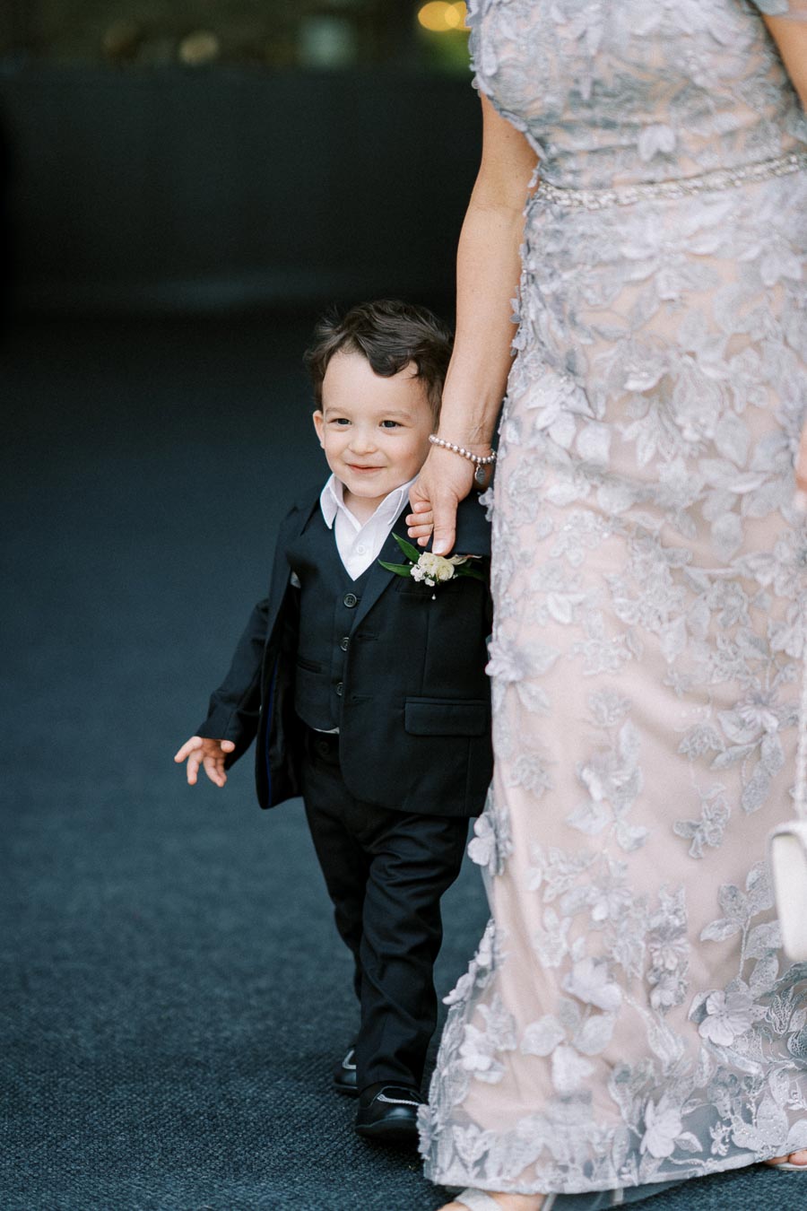 A young boy in a black suit with a boutonniere holding an adult's hand, walking at a formal event, accompanied by a person in a floral embroidered gown.
