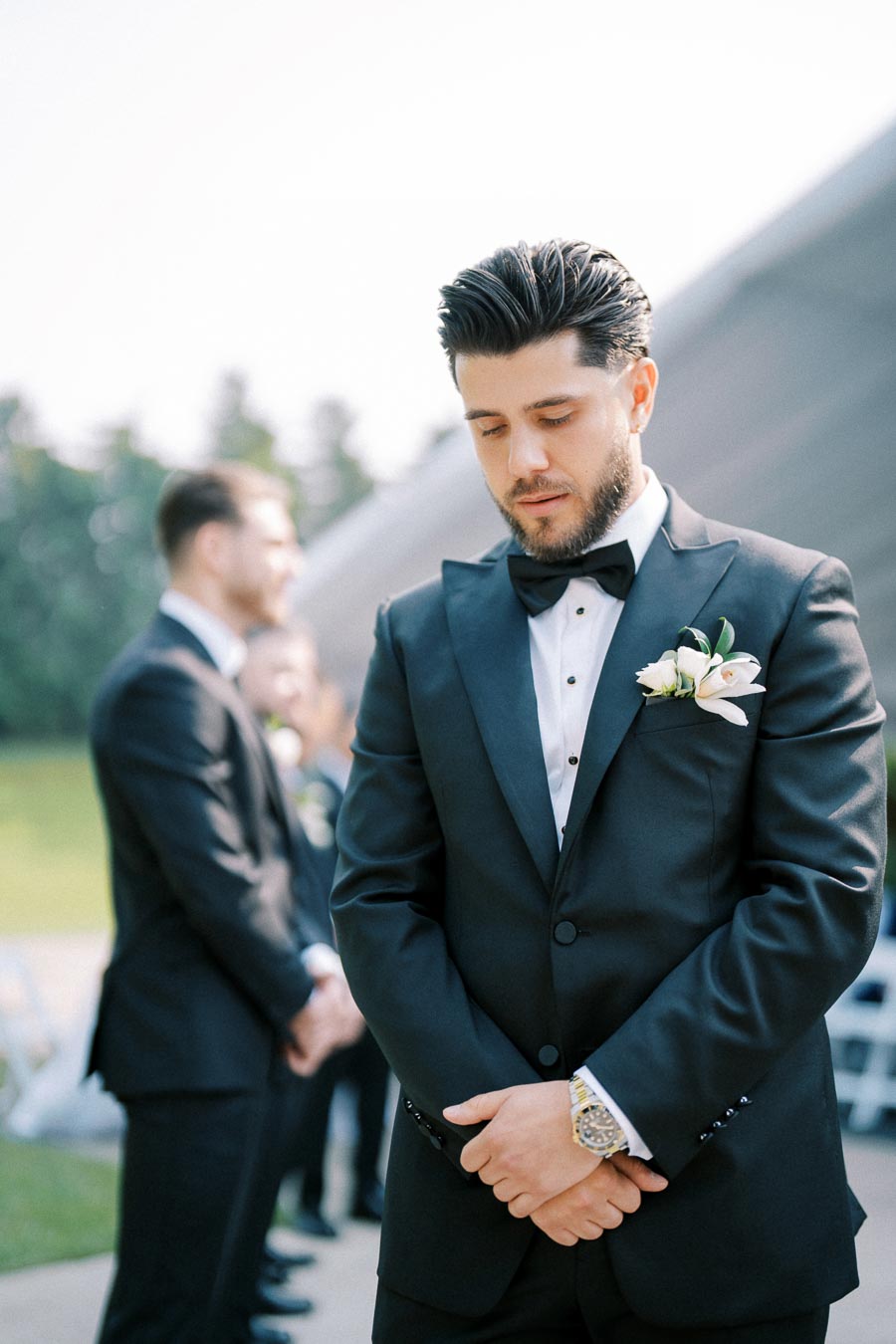 A groom in a black tuxedo with a bow tie and boutonniere stands pensively outdoors at a wedding ceremony, alongside his groomsmen.