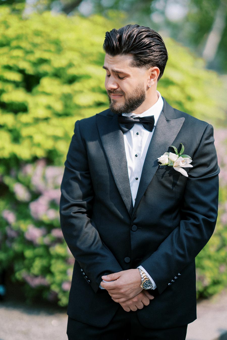 A groom in a black tuxedo with a white floral boutonniere standing outside against a background of lush greenery and purple flowers, looking to the side with a thoughtful expression.