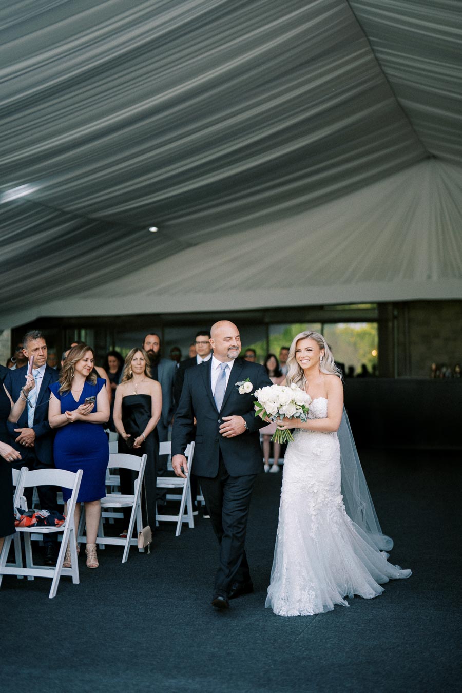 A bride in a white lace wedding dress and veil walks arm in arm with a man in a dark suit down the aisle of a wedding ceremony, surrounded by guests seated on white chairs inside a tent with draped ceilings.