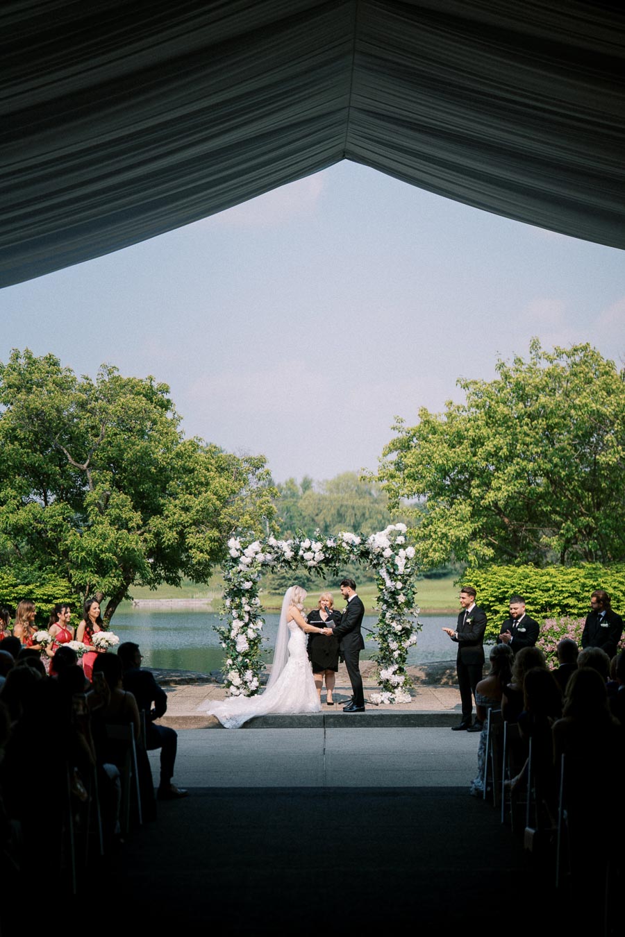 Outdoor wedding ceremony by a lake, featuring a bride and groom standing under a floral arch with guests seated on either side, surrounded by lush greenery.