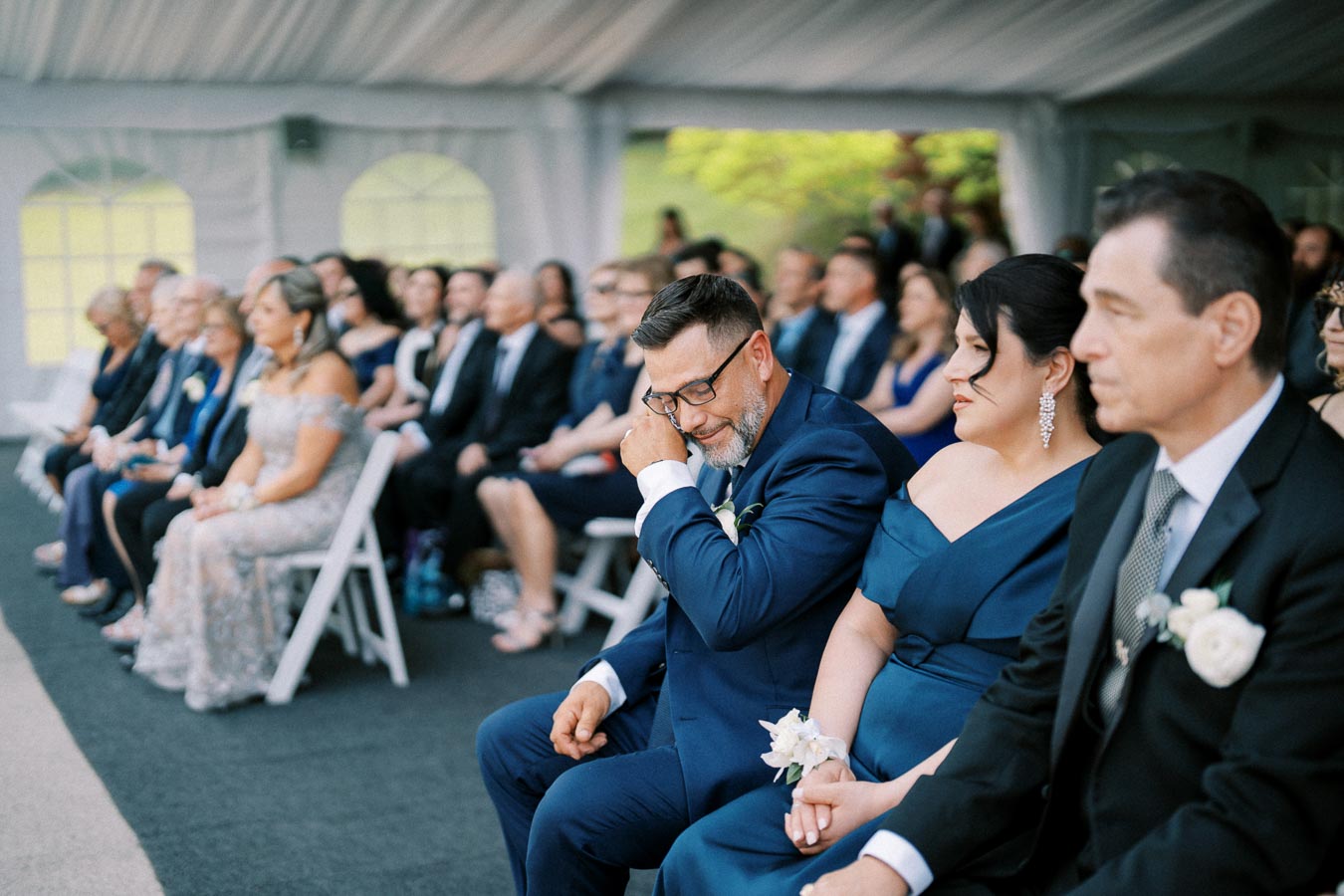 A wedding ceremony with a man in a blue suit wiping a tear, seated next to a woman in a blue dress, surrounded by guests, under a tent.