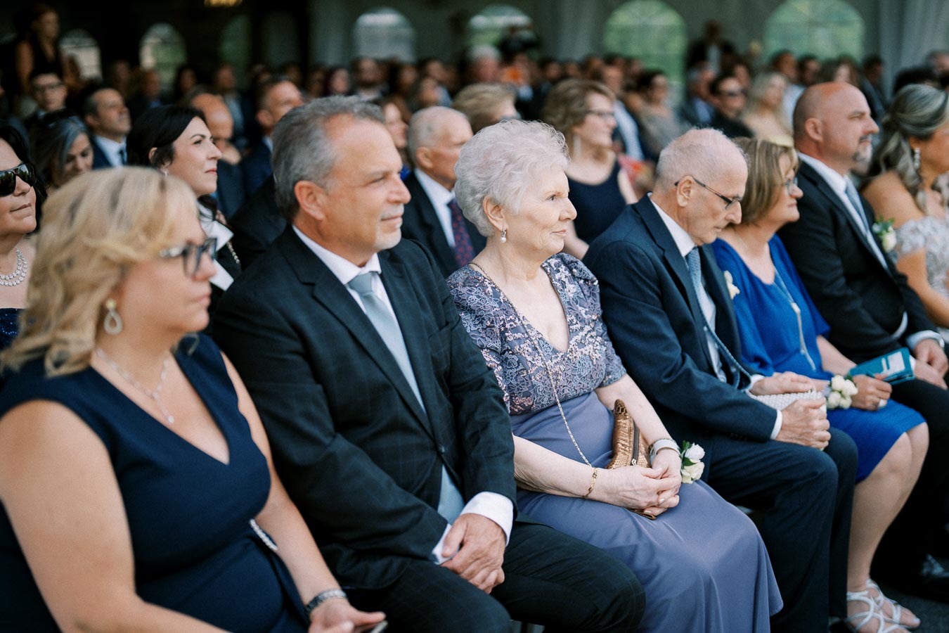 Group of people seated at an outdoor event, elegantly dressed in formal attire, attentively observing the proceedings.