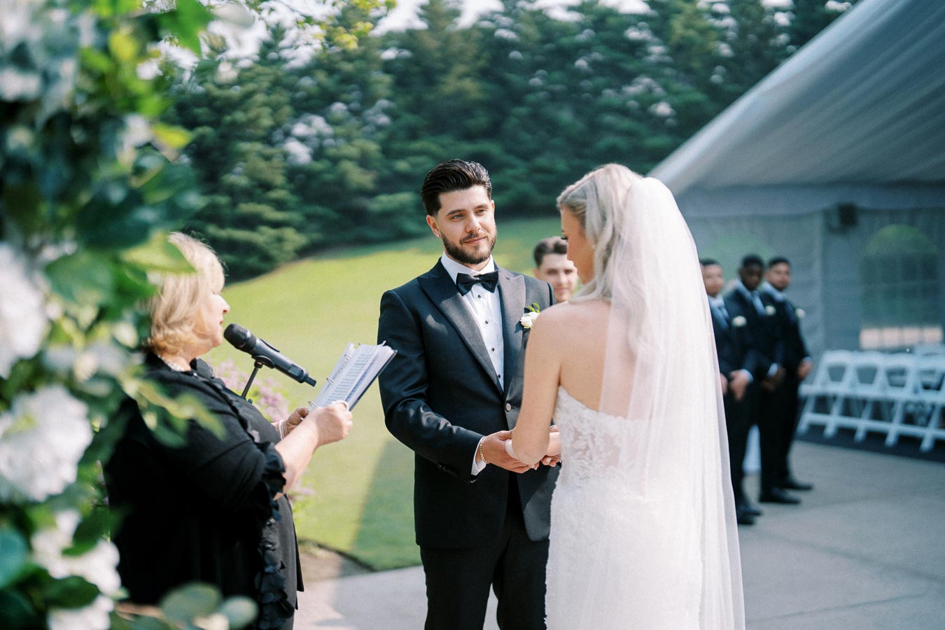 Outdoor wedding ceremony with bride and groom exchanging vows under a tent, surrounded by greenery.