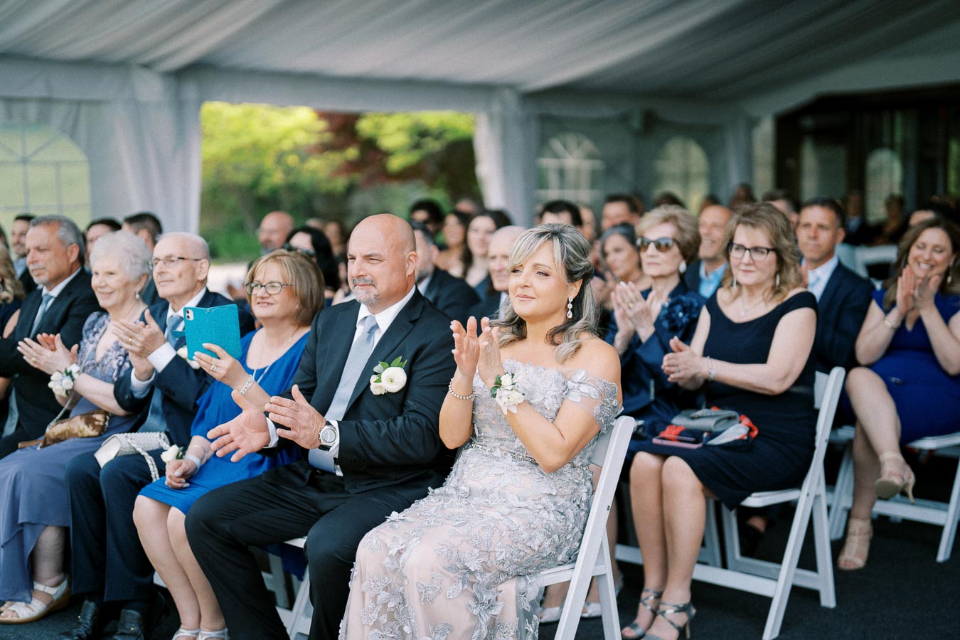 Guests in formal attire seated in a wedding ceremony under a tent, clapping and smiling, with a woman in an elegant silver dress and a man in a dark suit in the foreground.