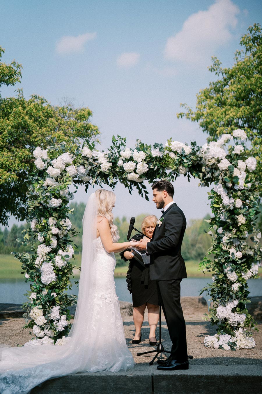 A couple exchanging vows under a floral wedding arch decorated with white flowers and greenery during an outdoor ceremony by a lake, with a clear blue sky overhead.
