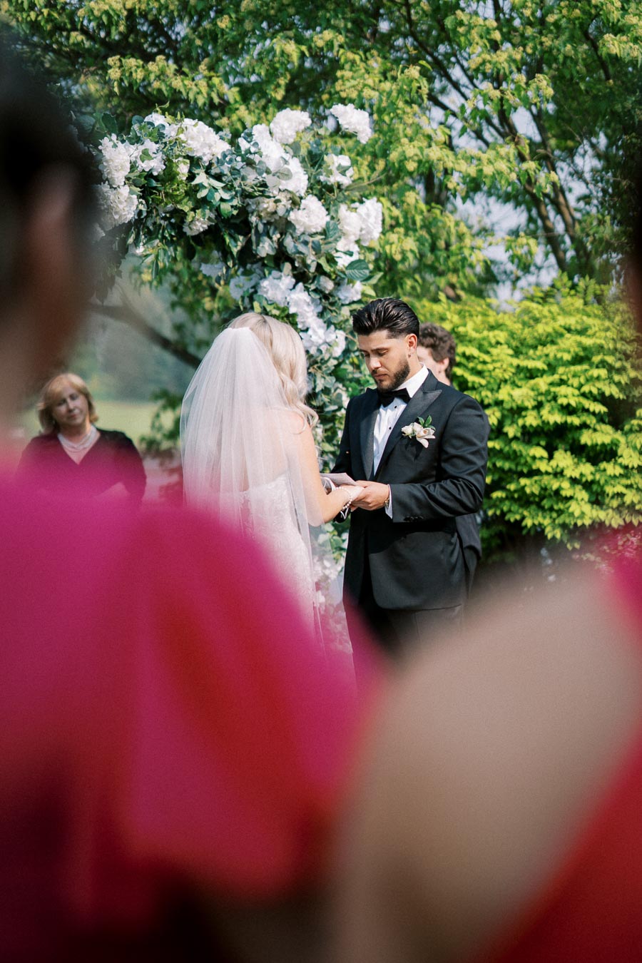 A bride and groom exchange vows under a floral arch during an outdoor wedding ceremony.