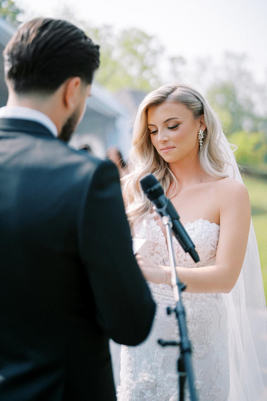 Bride and groom exchanging vows outdoors during a wedding ceremony, with the bride in a lace wedding dress and veil, and the groom in a black suit, captured on a sunny day.