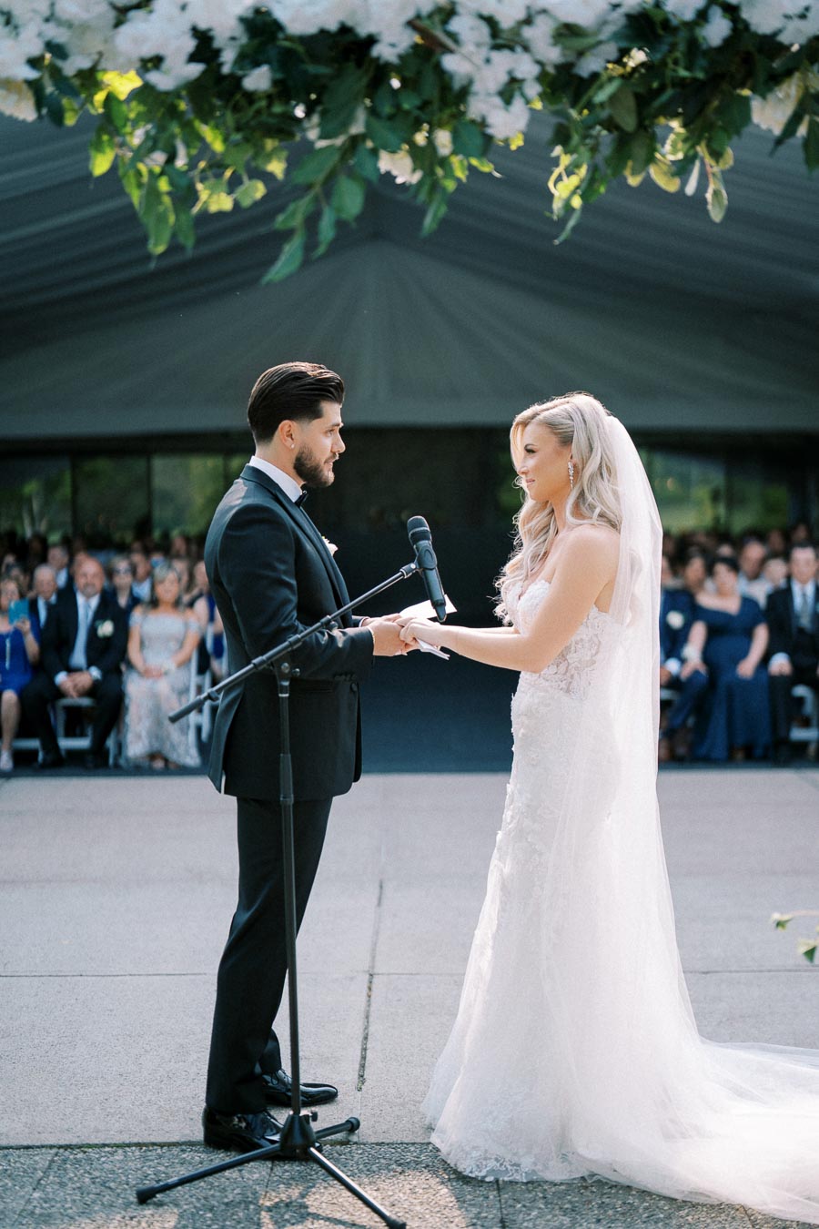 A bride and groom exchanging vows during an outdoor wedding ceremony, under a canopy of greenery, with guests seated in the background.