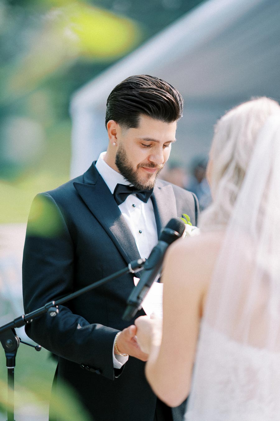 Groom in a black tuxedo sharing vows with bride during an outdoor wedding ceremony, with a microphone in the foreground and blurred greenery in the background.