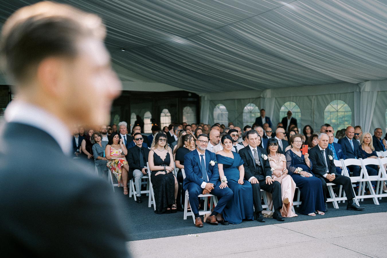 A blurred groom in the foreground with well-dressed guests seated under a decorated marquee tent at a wedding ceremony.