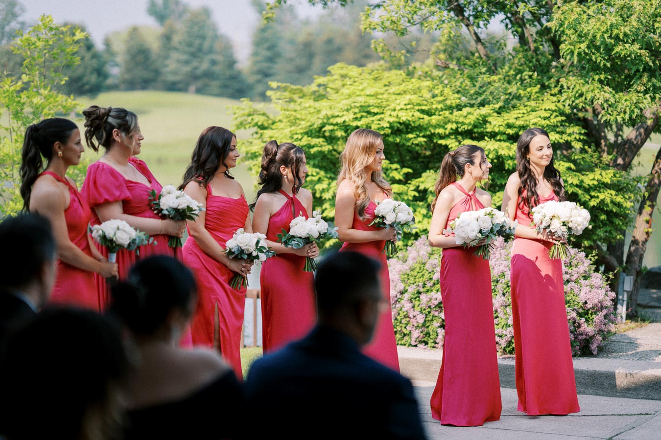 Bridesmaids in matching pink dresses holding white rose bouquets, standing outdoors in a lush green garden setting.