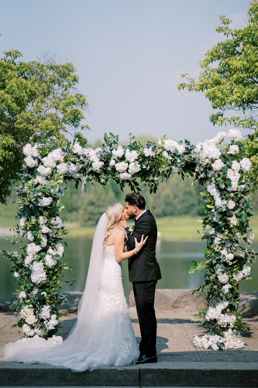 A bride and groom share a romantic kiss under a floral wedding arch adorned with white flowers and greenery, set against a picturesque outdoor backdrop with trees and a lake.
