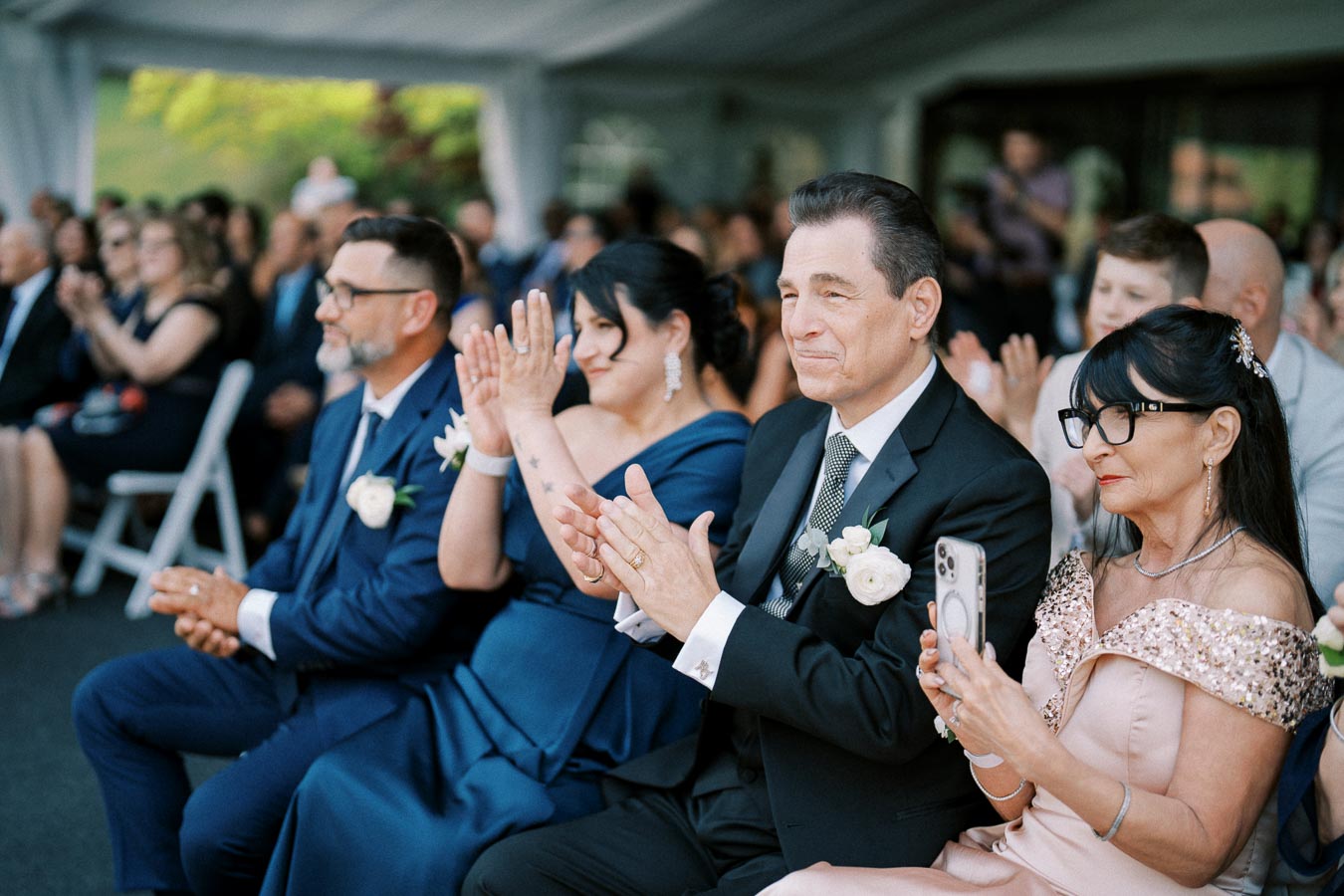 Guests in formal attire clapping during an outdoor wedding ceremony under a tent.