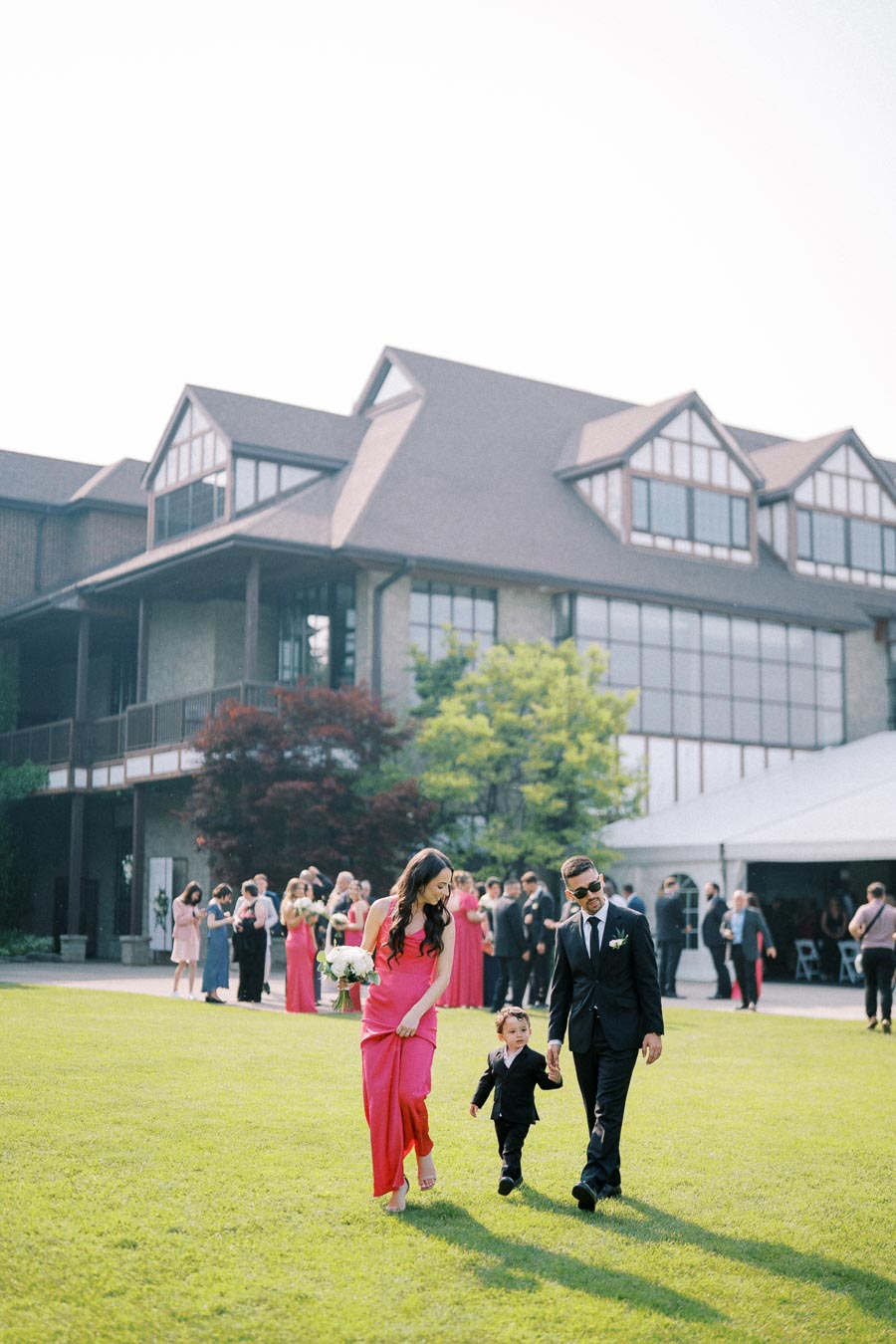 A stylish couple and a small child walk hand in hand on a lush green lawn during a formal outdoor event, with a large, elegant building and a group of well-dressed people in the background.