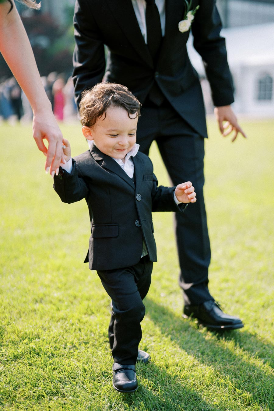 A toddler in a black suit happily holding hands with adults, walking on green grass during a sunny outdoor event.