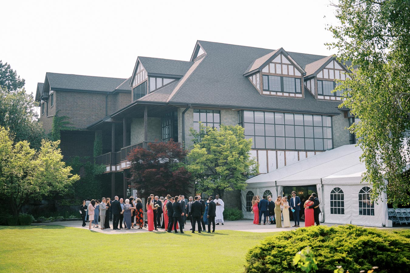 Outdoor wedding reception at a large, elegant estate with a crowd of guests mingling near a white tent and lush greenery surrounding the venue.