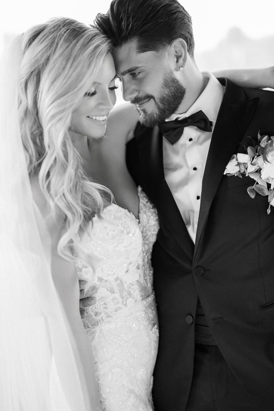 Black and white photo of a smiling bride and groom embracing on their wedding day, wearing elegant attire with the groom in a tuxedo and the bride in a lace wedding dress with veil.