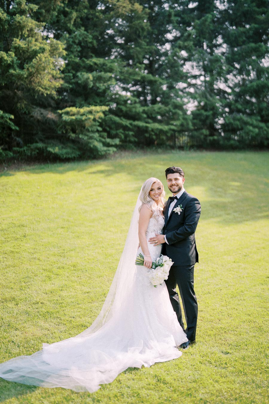 A bride in a lace wedding gown and veil stands with a groom in a black tuxedo, both smiling on a lush green lawn with evergreen trees in the background.