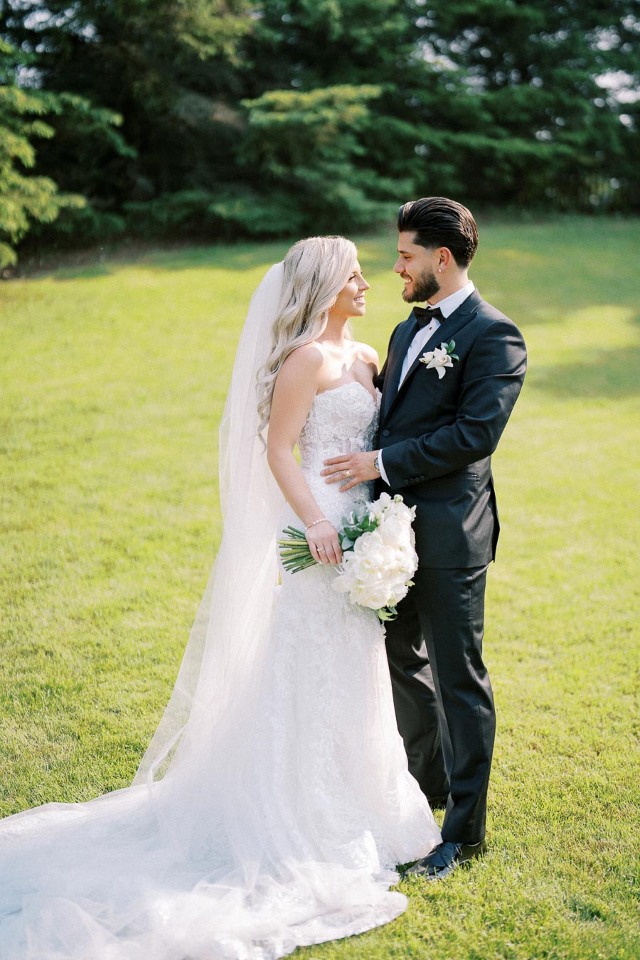 A bride and groom standing on lush green grass, gazing at each other lovingly, with the bride in a white lace gown holding a bouquet of white flowers, and the groom in a black tuxedo with a white boutonniere, set against a backdrop of vibrant greenery.
