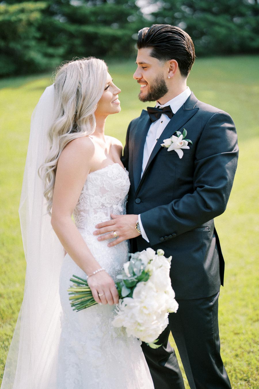 Bride and groom smiling at each other on their wedding day, the bride in a lace wedding dress holding a bouquet of white flowers, and the groom in a black tuxedo with a boutonniere, standing outdoors on green grass with trees in the background.