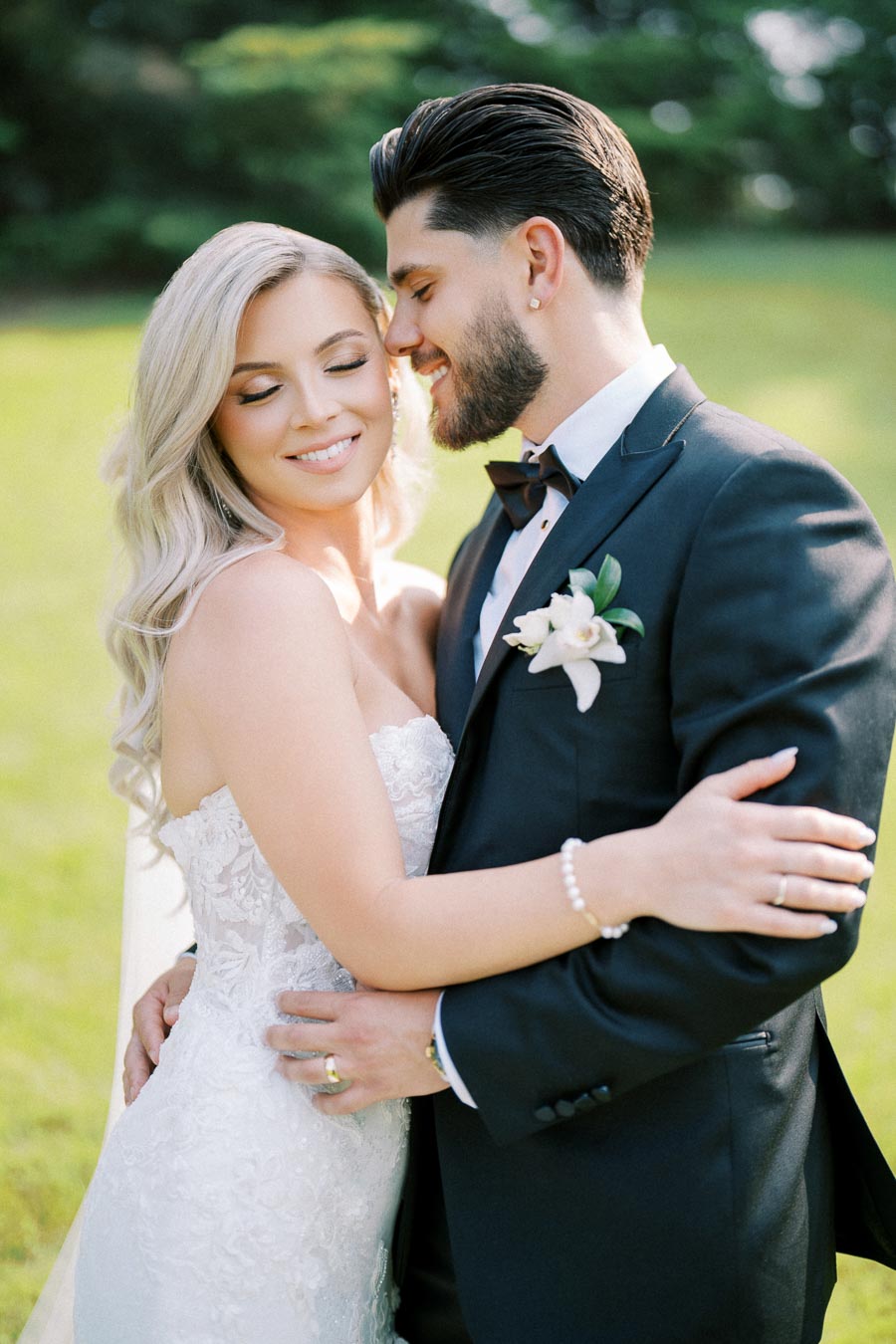 A bride and groom embracing in a sunlit garden, the bride wearing a lace wedding dress and the groom in a black tuxedo with a white boutonniere, both smiling blissfully on their wedding day.