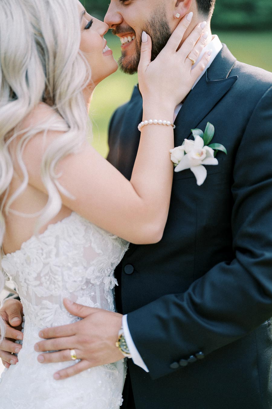 A bride and groom lovingly embrace on their wedding day, with the bride wearing a lace gown and the groom in a dark suit adorned with a floral boutonniere.