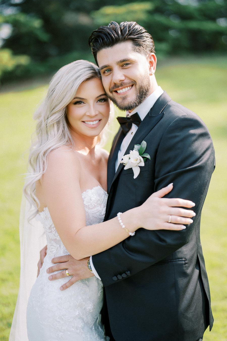 Smiling couple in wedding attire embracing outdoors, featuring a bride in a lace gown and a groom in a black tuxedo with a floral boutonniere, standing against a lush green background.