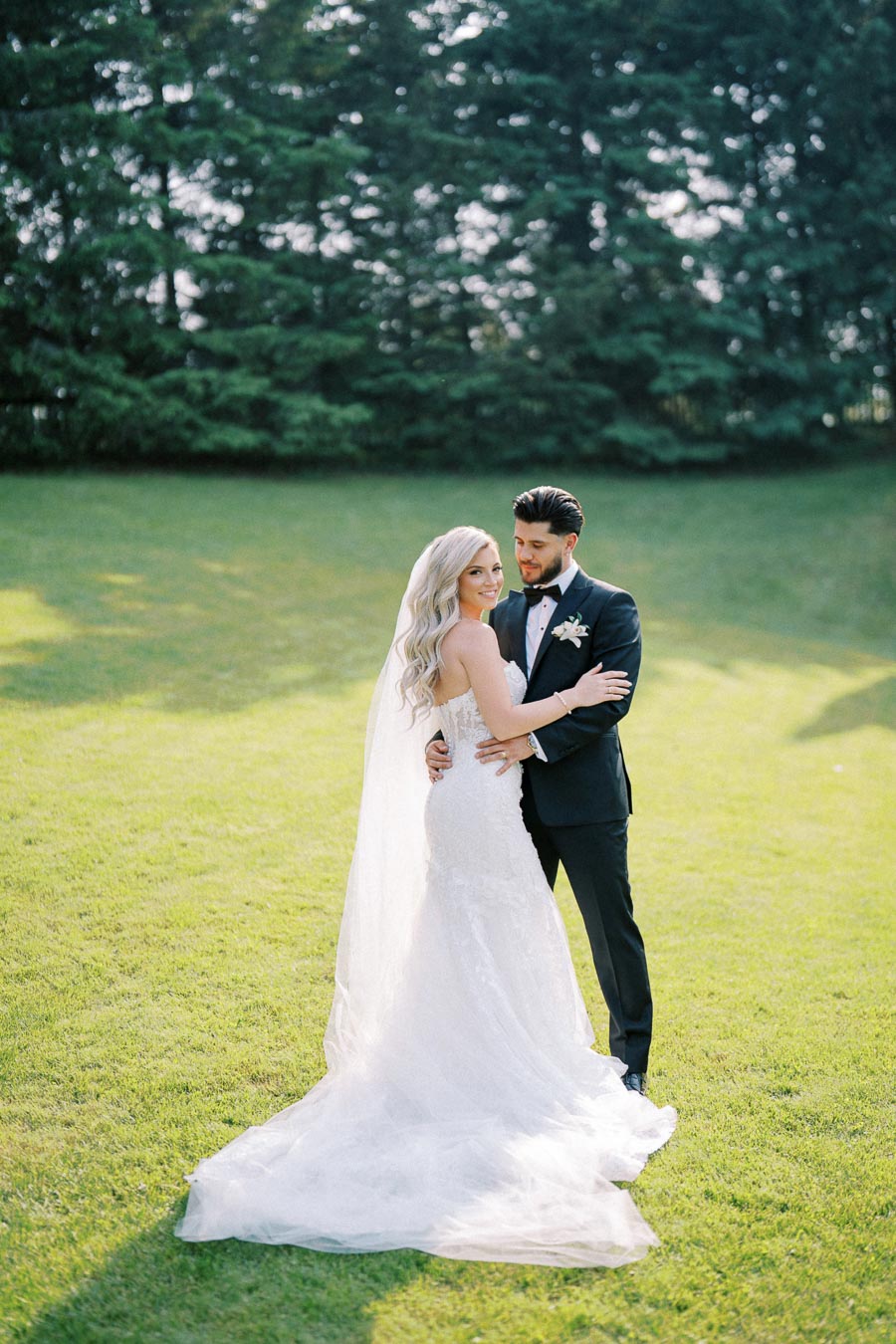 Elegant bride and groom embracing in a sunny garden on their wedding day, with a lush green background and the bride wearing a flowing white gown and veil.
